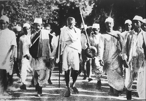 A photograph of Mohandas Gandhi is shown. There are several people walking with him.