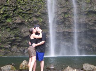 Photograph shows a couple embracing and kissing next to a waterfall.