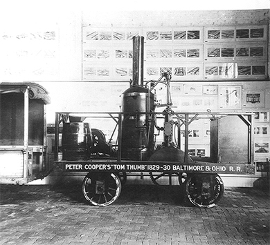 A photograph of a replica of the Tom Thumb steam locomotive is shown. On its side are painted the words “Peter Cooper’s ‘Tom Thumb’ 1829–30 Baltimore and Ohio R.R.”