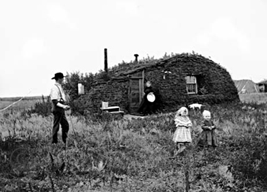 A photograph of a sod hut is shown. Before it stand a farmer, his wife, and two children.