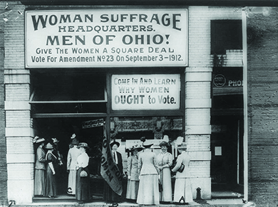 A photograph shows women suffragists standing outside a building. The sign above them reads "Woman Suffrage Headquarters. Men of Ohio! Give The Women A Square Deal. Vote For Amendment No. 23 on September 3—1912." A second sign reads "Come In And Learn Why Women OUGHT to Vote."