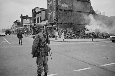 A photograph shows a street, deserted but for one pedestrian and several men in riot gear. The ruins of a building are visible on the corner.