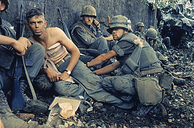A photograph shows a group of uniformed U.S. soldiers crouched beside a wall. One soldier is shirtless, with a large bandage wrapped around his chest.