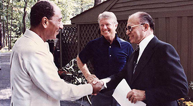 A photograph shows Jimmy Carter standing by as Anwar Sadat shakes hands with Menachem Begin.