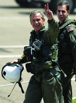 A photograph shows George W. Bush walking with naval flight officer Lt. Ryan Phillips following the president’s arrival on the USS Abraham Lincoln. Bush wears a flight suit and gives a victory symbol to the cameras with his hand.