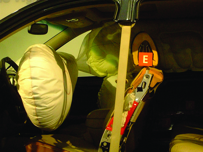 This photograph shows the inside of an automobile from the driver’s side area. The image shows inflated airbags positioned just in front of the driver’s and passenger’s seats and along the length of the passenger side over the windows. A large, round airbag covers the steering wheel.