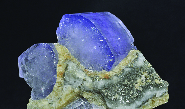 This figure includes an image of two large light blue apatite crystals in a mineral conglomerate that includes white, grey, and tan crystals. The blue apatite crystals have a dull, dusty, or powdered appearance.