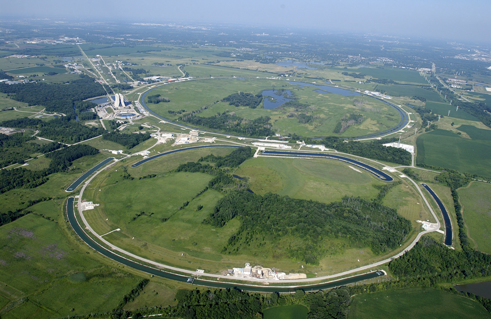 An aerial view of the Fermi National Accelerator Laboratory. The accelerator has two large, ring shaped structures. There are circular ponds near the rings.
