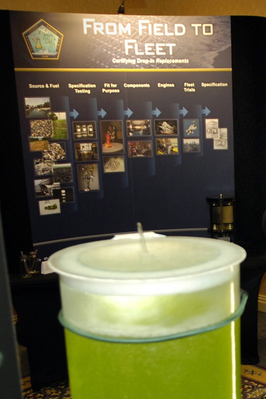 Image of a poster in front of a barrel of algae producing biofuel.