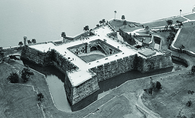 An aerial photograph shows the Spanish fort of Castillo de San Marcos, a square, high-walled structure facing the water and including a surrounding moat.