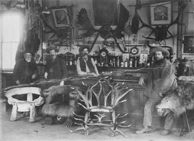 A photograph shows the interior of a saloon. Several chairs constructed of wood, hides, and antlers sit in front of a bar. Several customers sit or stand at the bar. Another man stands behind the bar, along with bottles of liquor, several framed images, and a variety of decorations and wall hangings.