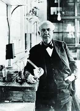 A photograph shows Thomas Edison in a brightly lit workroom. Beside him is a table holding an incandescent light bulb.