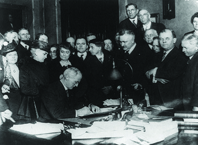A photograph depicts Governor James P. Goodrich signing a bill, surrounded by a large group of men and women.