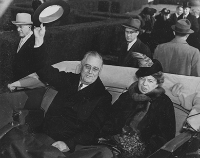 A photograph shows Franklin and Eleanor Roosevelt smiling as they ride in the back of a coach. Franklin Roosevelt waves his hat at onlookers.