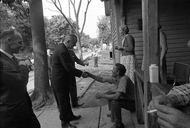 A photograph shows President Johnson standing on a street outside of a house, several of whose inhabitants sit and stand on the porch. He shakes the hand of a seated man while two other officials look on.