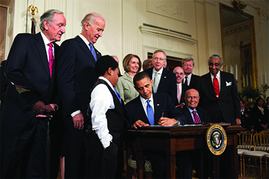 A photograph shows President Obama signing the Patient Protection and Affordable Care Act as Vice President Biden, Speaker of the House Nancy Pelosi, Senate Majority Leader Harry Reid, and others look on.